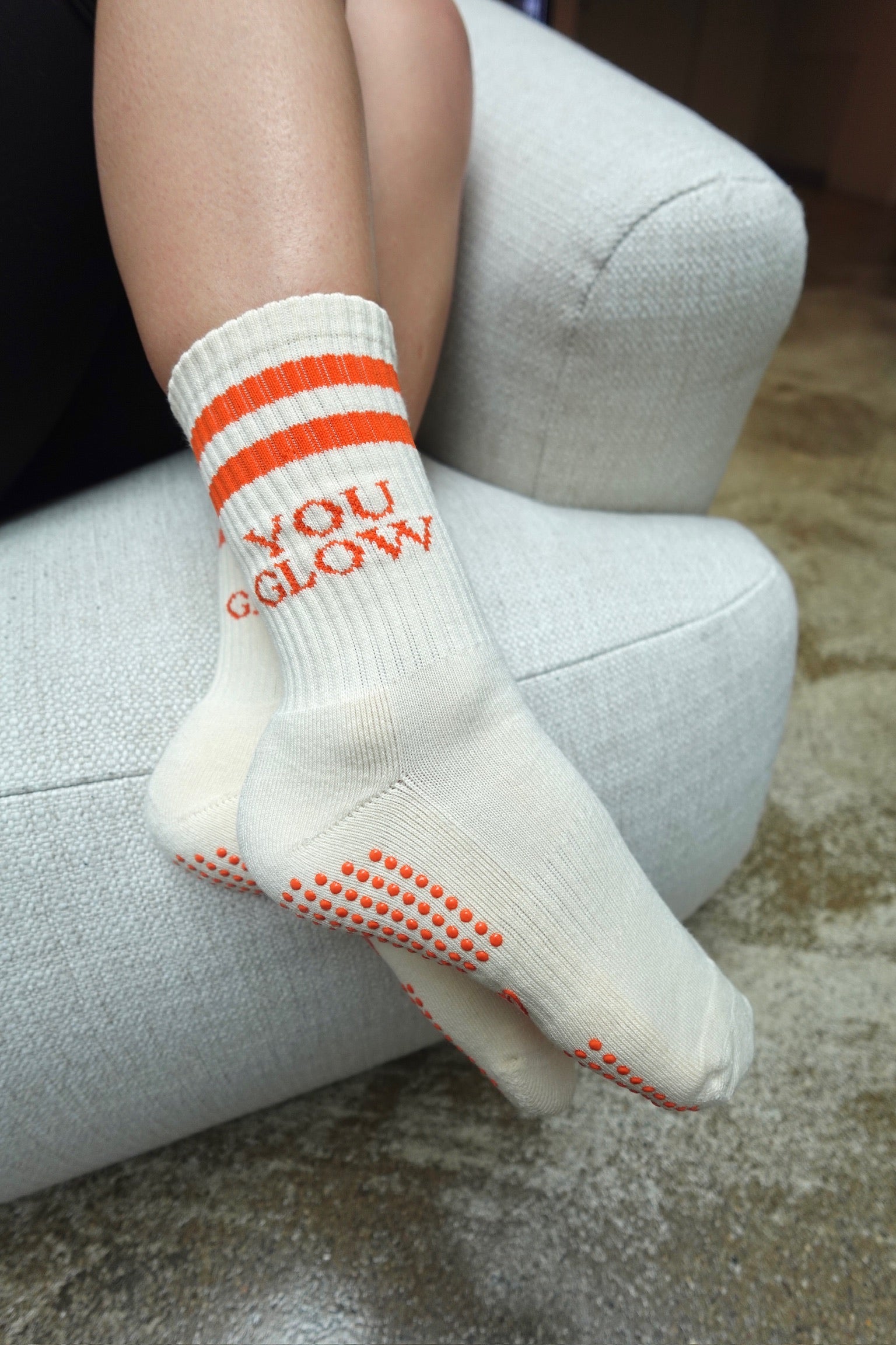 Cremefarbene Pilates-Socken „You Glow“ mit orangefarbenen Streifen und rutschfesten Punkten, liegend auf Stoffoberfläche