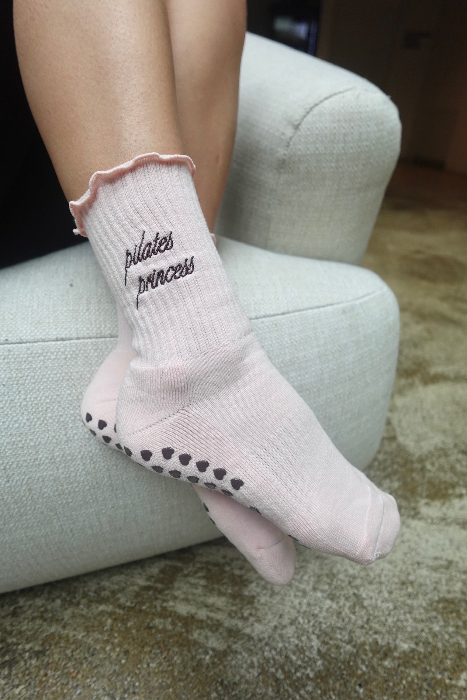 Rosa Pilates-Socken „Pilates Princess“ mit dunkler Stickerei und Herz-Anti-Rutsch-Noppen, getragen auf hellem Sessel